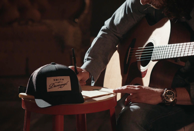 Smith Devereux Merlot Showcase Close-up of a musician writing lyrics on a notepad while holding an acoustic guitar, with a Smith Devereux branded hat on a stool nearby.