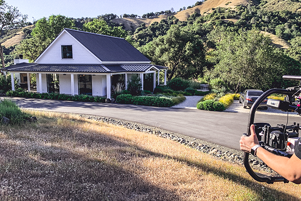 Promise Wine Still 2 A person is holding a camera rig, filming a white house with a black roof in a scenic, hilly area with trees and greenery.