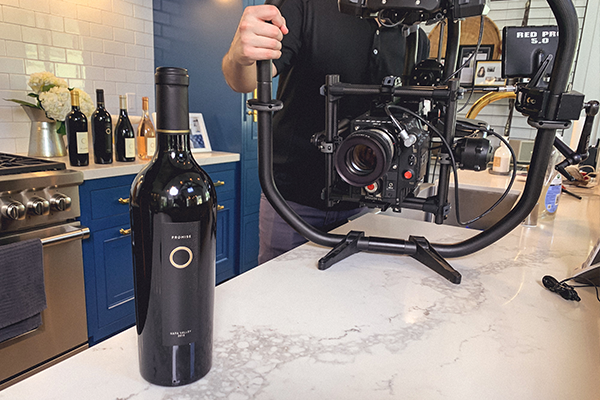 Promise Wine Still 3 A person is holding a camera rig on a kitchen counter. In the foreground, there is a bottle of wine.