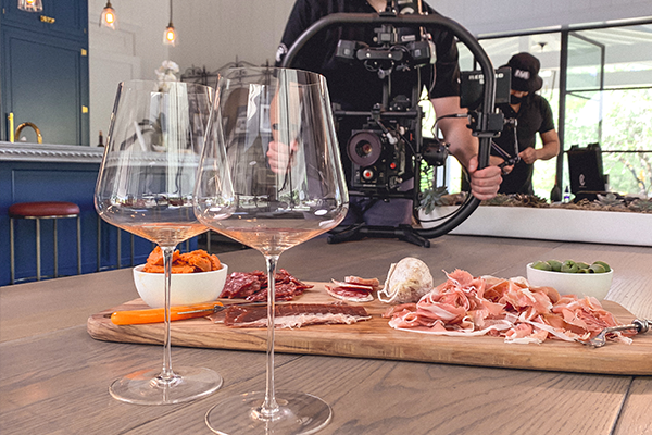 Promise Wine Still 5 A close-up of a charcuterie board along with two wine glasses on a wooden table. In the background, a person operates a camera rig.
