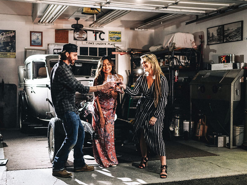 Smith Devereux Garage Tasting Three people standing in a garage with a vintage car in the background, sharing a drink.