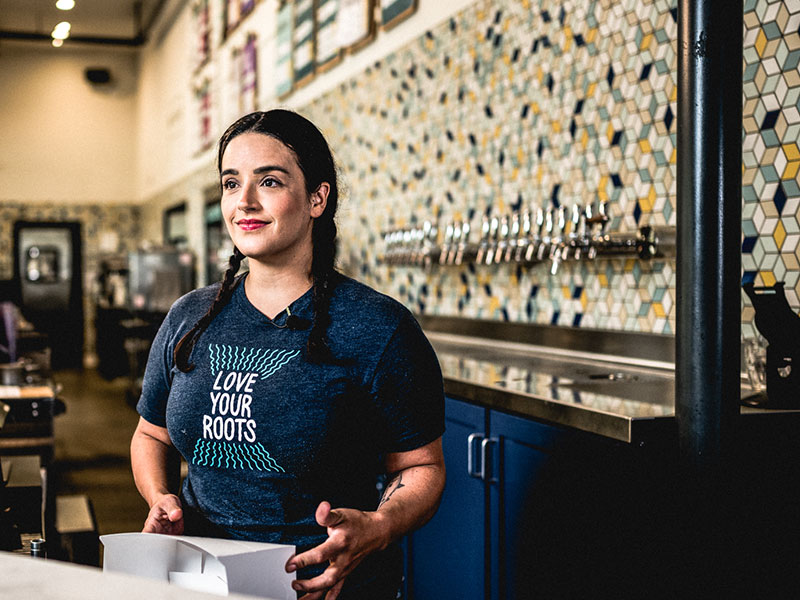 urban roots beer actress on video A woman in a bar with shelves of liquor bottles behind her.