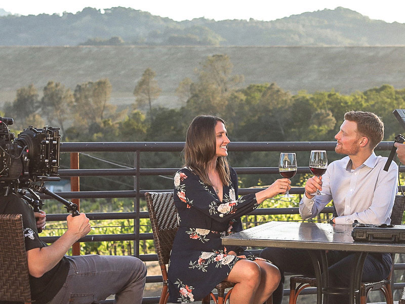 A camera crew is filming two people sitting at an outdoor table on a balcony, toasting with glasses of red wine.