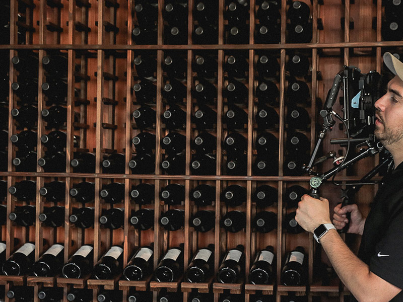 Behind-the-scenes of Pour's crew member holding a camera rig, filming in front of a wine rack filled with numerous bottles of wine.