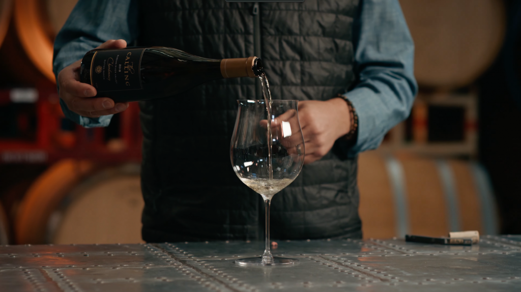 A person is pouring white wine from a bottle labeled 'The Calling' into a wine glass on a metal table.
